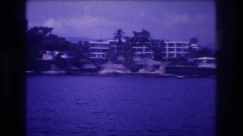 Clearest 8mm frame showing a large multi-story building with horizontal balcony tiers nestled in tropical vegetation above a rocky shoreline, Acapulco area circa 1982.