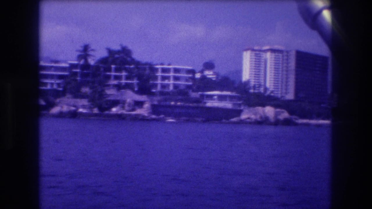 1982 Acapulco, Mexico: 8mm home-movie view from a moving boat toward a rocky headland lined with palm trees and high-rise resort hotels, heavy blue-purple film shift.