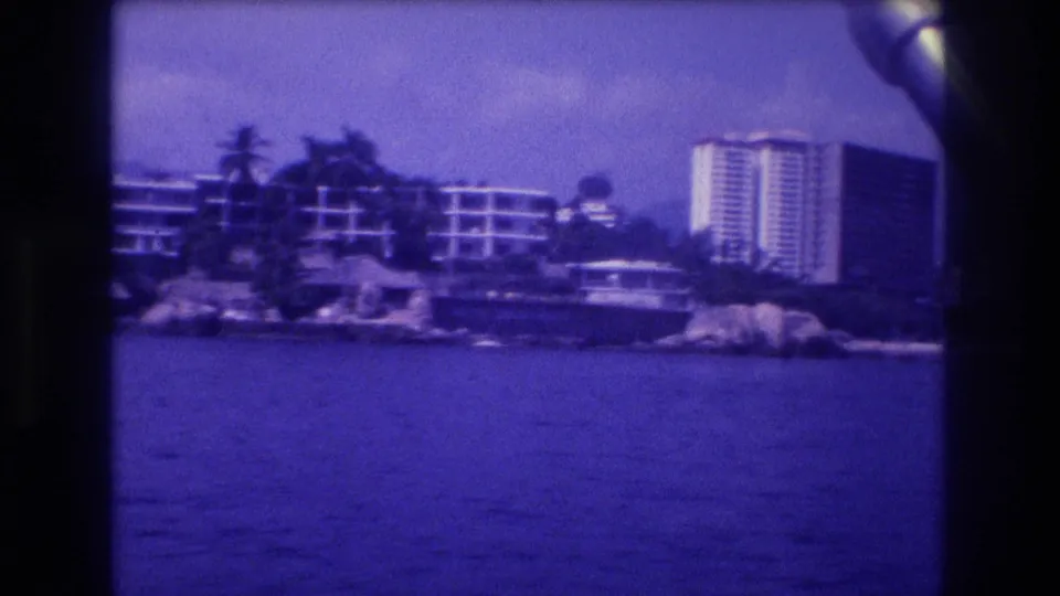 1982 Acapulco, Mexico: 8mm home-movie view from a moving boat toward a rocky headland lined with palm trees and high-rise resort hotels, heavy blue-purple film shift.