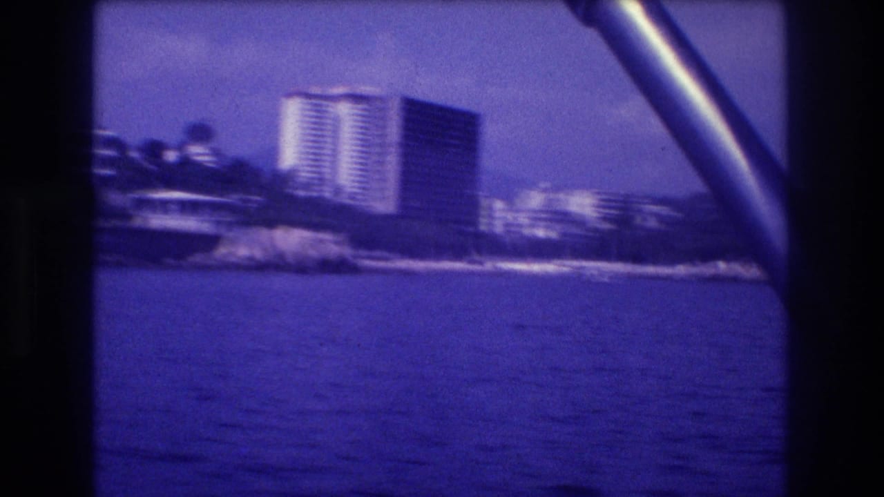 Two tall white high-rise buildings on a tropical headland seen from a boat, with water in the foreground and a metal boat rail at frame edge.