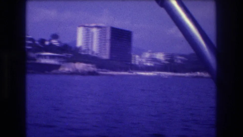 Acapulco bay area, 1982: two high-rise resort towers on a rocky palm-lined promontory, shot from a moving boat on blue-purple-cast 8mm film.