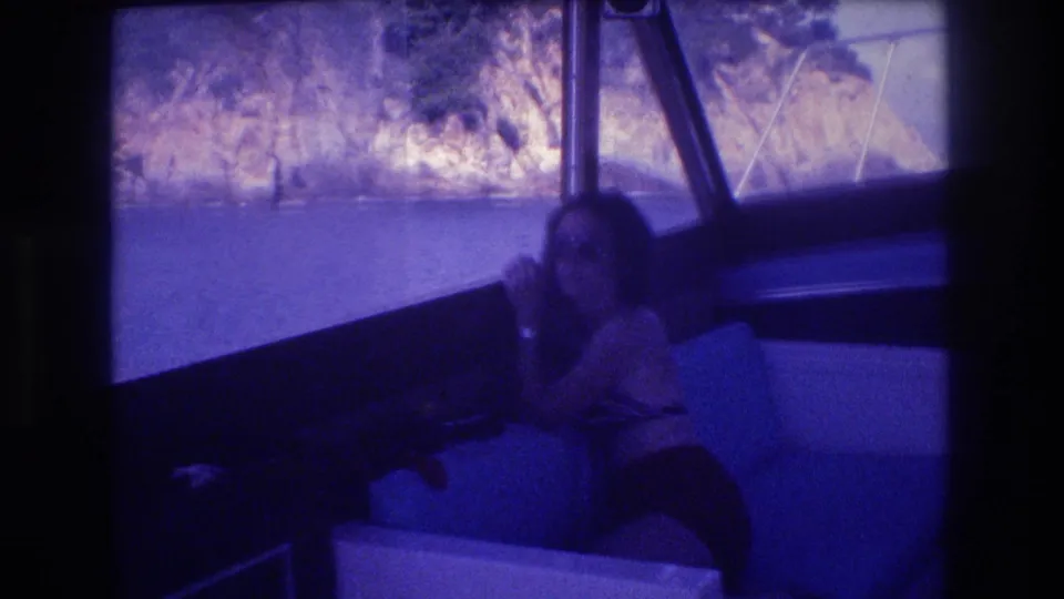 Acapulco boat trip, 1982: a woman relaxing on cushioned boat seating, tropical cliff vegetation behind her, captured on aged 8mm with blue-purple color shift.