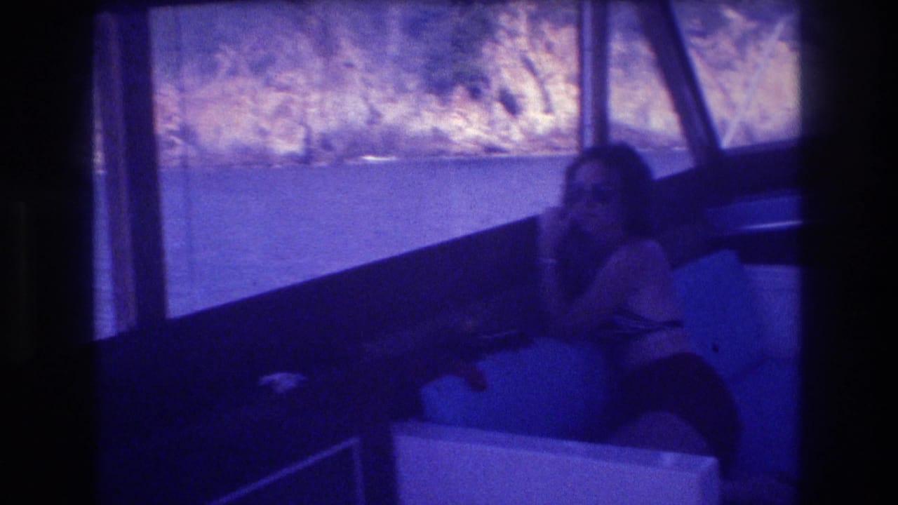 Woman in dark swimwear seated on blue upholstered bench inside a boat cabin, rocky coastal terrain visible through windows behind her.