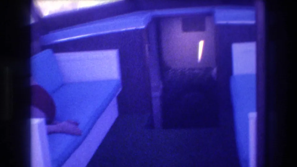 Interior of boat cabin, possibly Acapulco 1982, showing cushioned bench seating along the walls; a partial figure is visible at the lower left; warm interior lamp and heavy blue 8mm tint.
