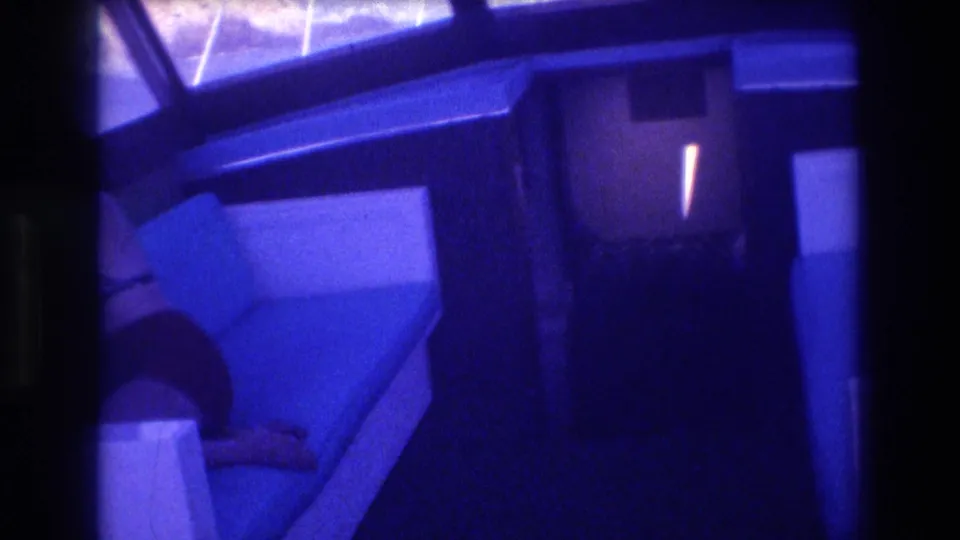 Boat cabin interior, possibly Acapulco 1982; blue bench seating with a partially visible dark-clothed figure at lower right; rocky coastal exterior through windows and heavy 8mm color shift.