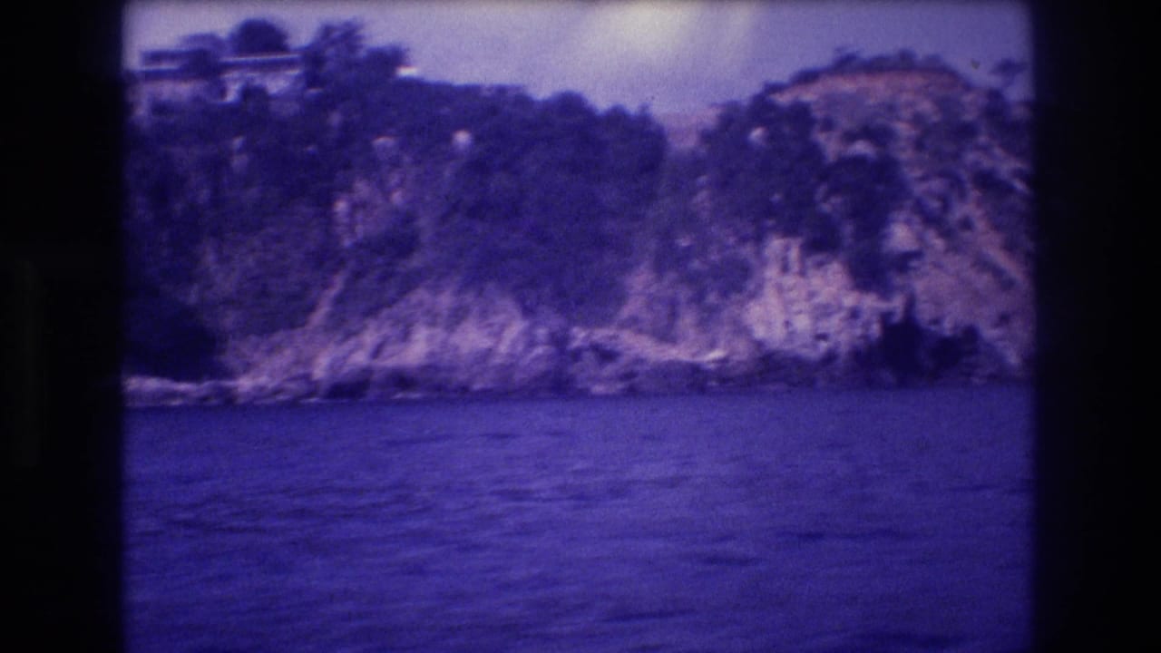 Rocky vegetation-covered headland viewed from a boat, deep blue-purple water below, hazy sky above.