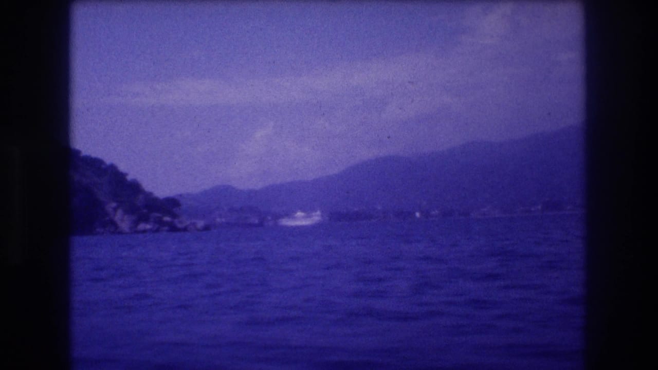8mm frame from a moving boat on Acapulco bay, Mexico, 1982, showing a receding rocky headland, hazy mountain ridgeline, and a small white motorboat in the distance.