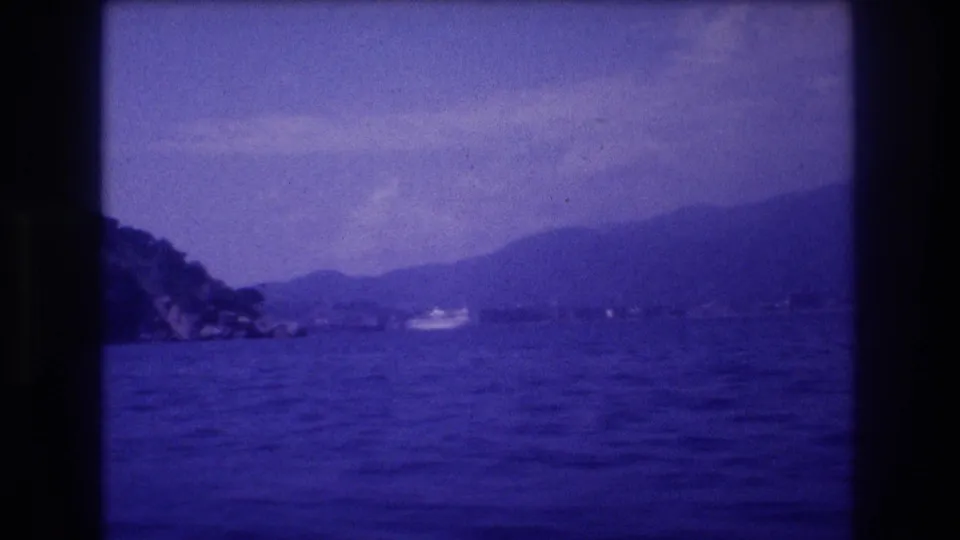 8mm frame from a moving boat on Acapulco bay, Mexico, 1982, showing a receding rocky headland, hazy mountain ridgeline, and a small white motorboat in the distance.