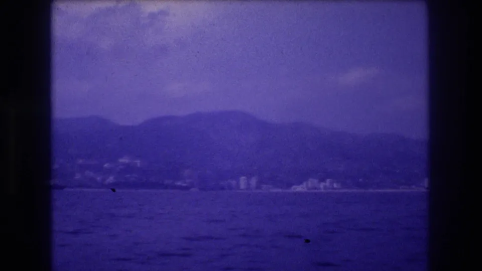 8mm home movie frame circa 1982 showing a developed coastline with buildings at the mountain base and ocean in the foreground, likely Acapulco, Mexico.
