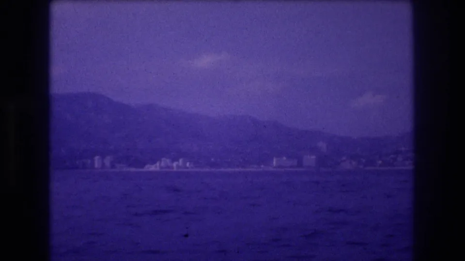 8mm home movie frame circa 1982 of a coastal resort city with multi-story buildings at the foot of mountains and ocean in the foreground, likely Acapulco, Mexico.