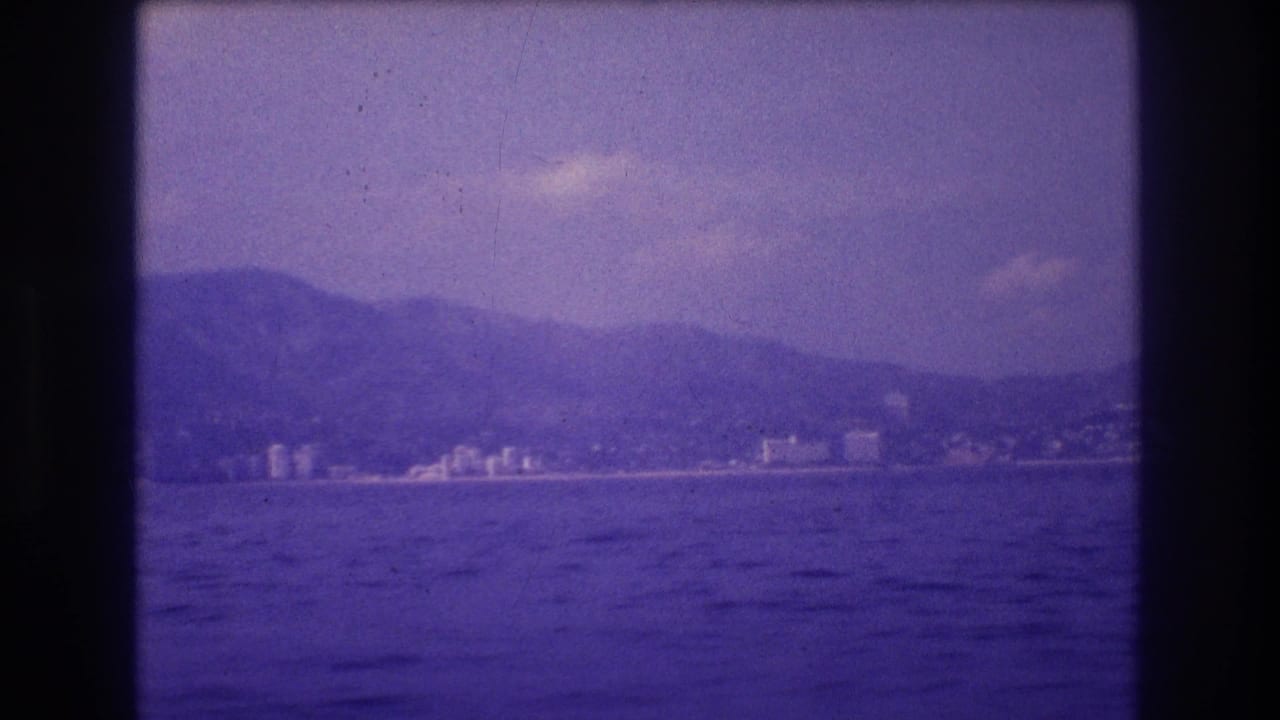 8mm home movie frame circa 1982 showing Acapulco's coastal skyline at distance from a boat on the Pacific, mountains in background, heavy purple-blue film cast.