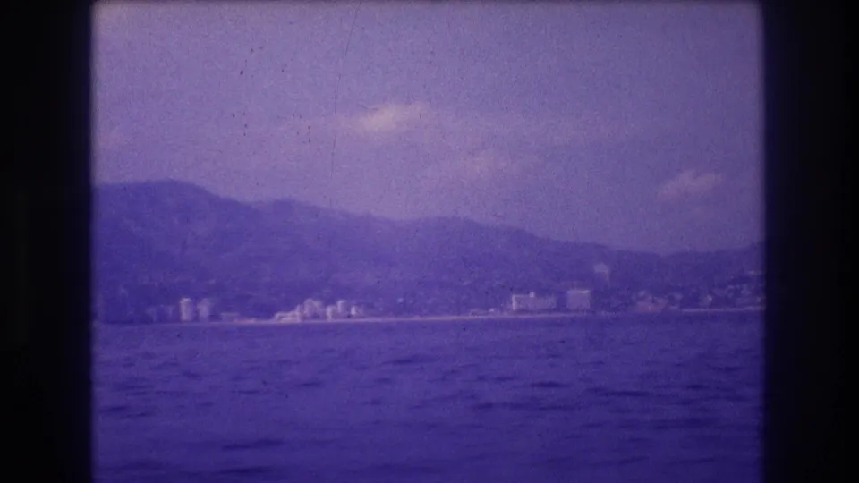 8mm home movie frame circa 1982 showing Acapulco's coastal skyline at distance from a boat on the Pacific, mountains in background, heavy purple-blue film cast.