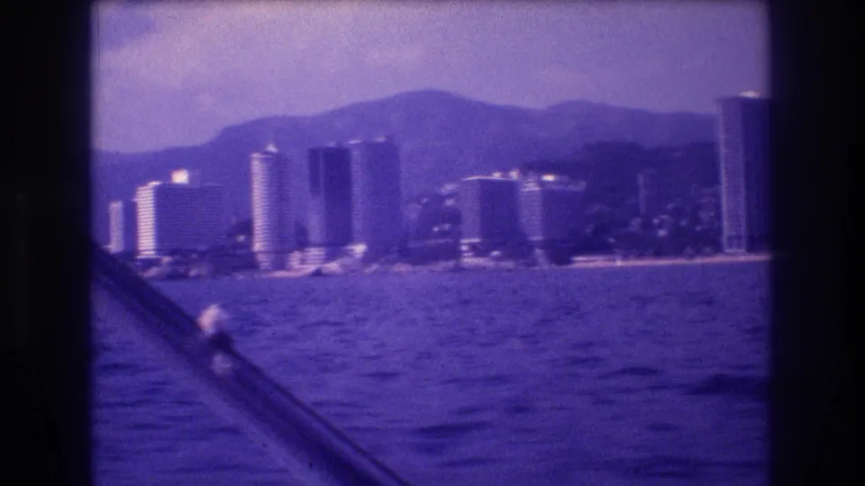 8mm home movie frame circa 1982 showing Acapulco's hotel tower strip from a boat, multiple tall buildings, mountain backdrop, boat railing foreground, purple-blue cast.