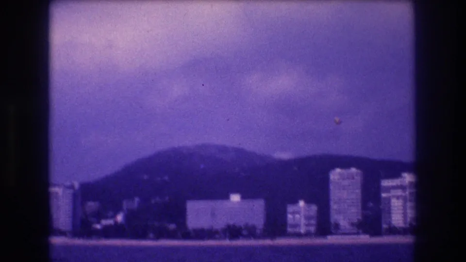 8mm home movie frame circa 1982 showing a stretch of Acapulco's bay coast with lower buildings and a mountain backdrop, purple-blue film cast throughout.