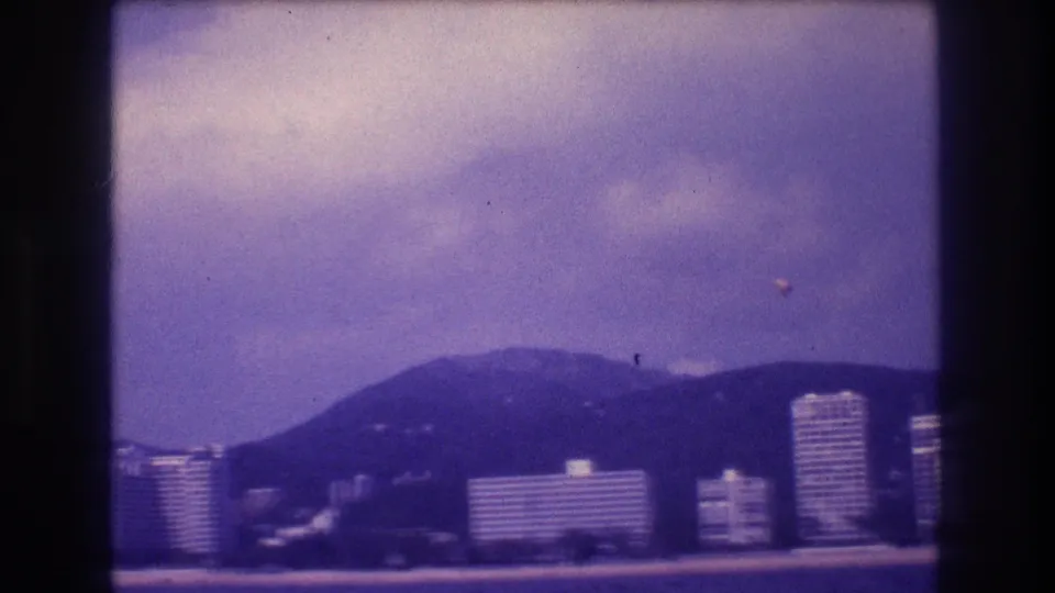 Aged 8mm coastal frame from Acapulco, Mexico, circa 1982: hotel towers line the bay, mountains rise behind, and a small distant object appears in the violet-shifted sky