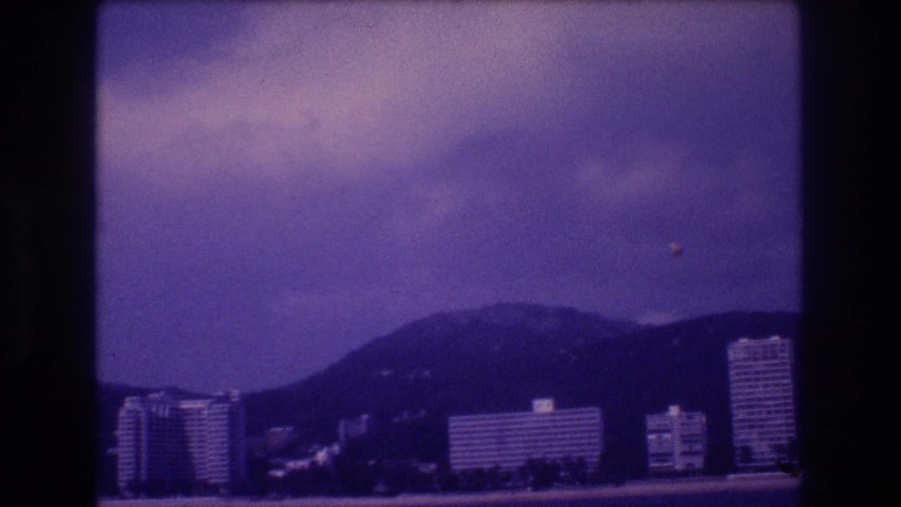 Acapulco coastal scene circa 1982 on aged 8mm; hotel towers line the bay with mountains behind and a small airborne speck visible in the upper-right sky
