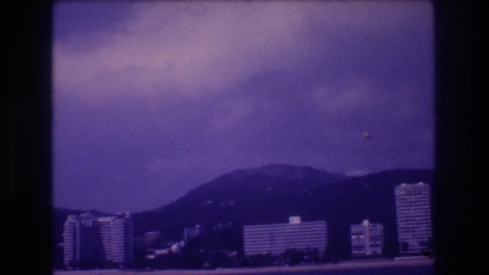 Acapulco coastal scene circa 1982 on aged 8mm; hotel towers line the bay with mountains behind and a small airborne speck visible in the upper-right sky
