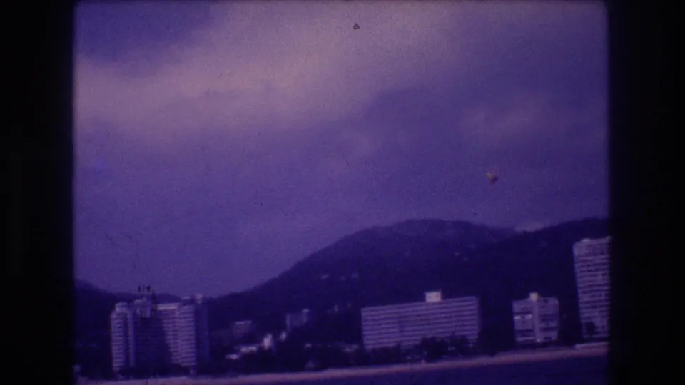 Camera tilting skyward in Acapulco, circa 1982; purple color-shifted 8mm frame shows overcast clouds above coastal hotel towers and hill line