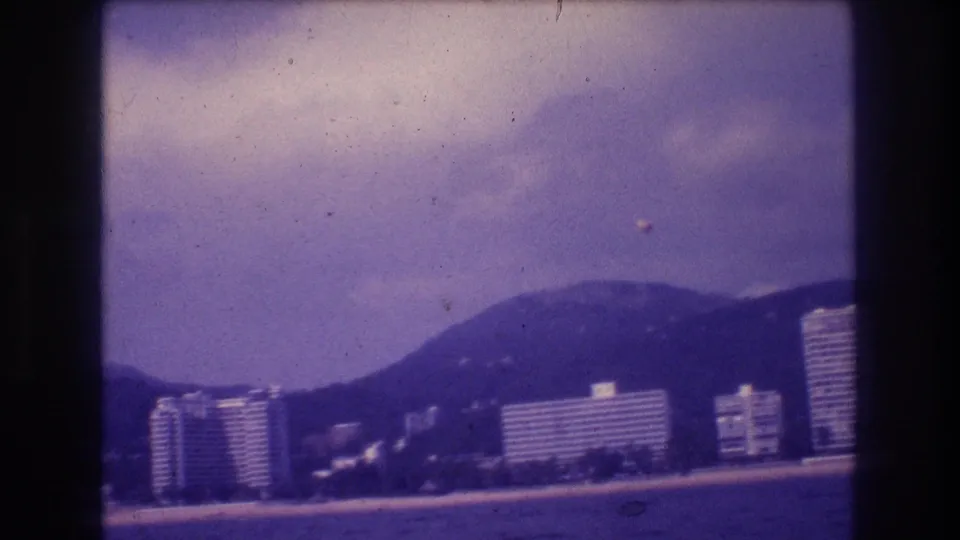 Final frame from 1982 Acapulco 8mm sequence; overcast sky dominates the purple-tinted image with coastal resort towers and mountain ridge at right
