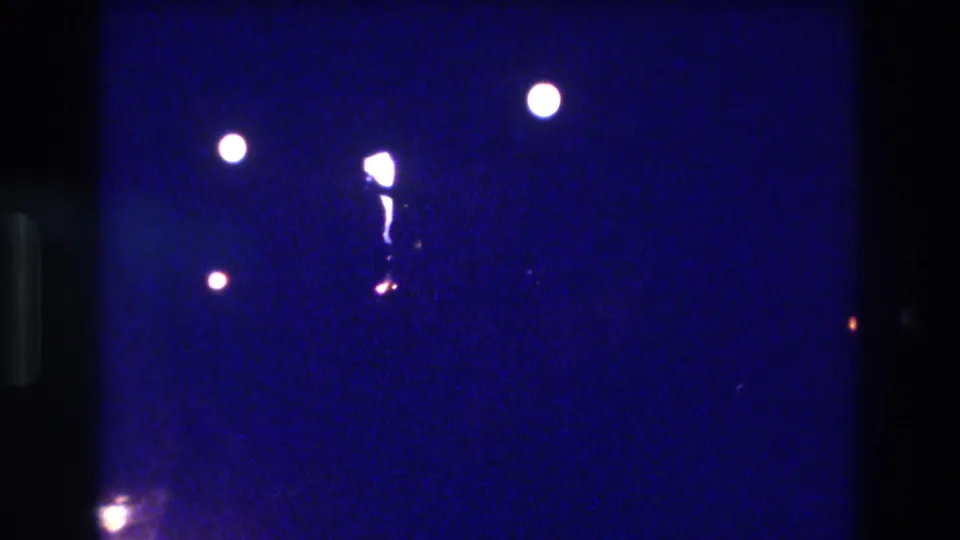 Circa 1982 8mm nighttime home movie frame showing three scattered white light orbs, a glowing elongated central shape, and a small warm-toned point light against heavy blue grain.