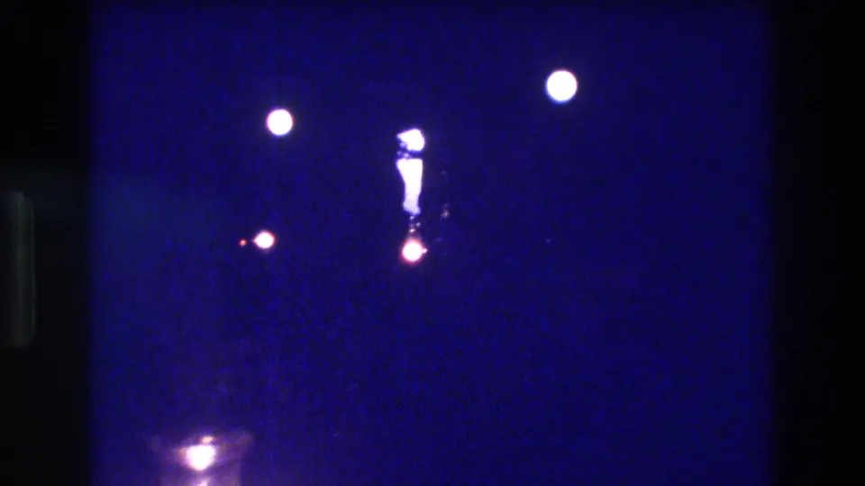Circa 1982 8mm nighttime home movie frame showing scattered light sources and a central glowing elongated form against heavy deep blue film grain; Acapulco per metadata.