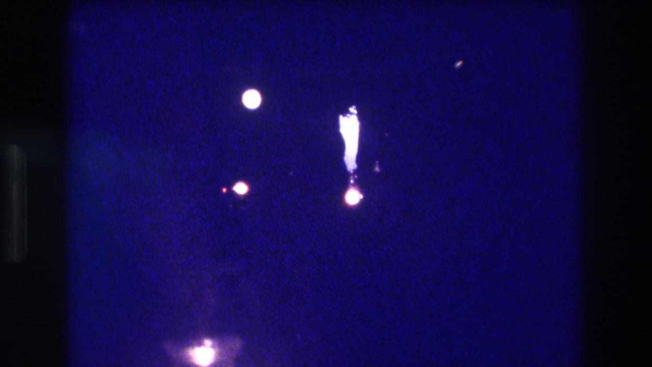 8mm home-movie frame from an Acapulco nighttime performance, circa 1982, showing spotlight orbs, a possible lit element or figure, and a bottom-of-frame glow against a saturated indigo field.