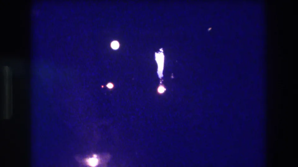 8mm home-movie frame from an Acapulco nighttime performance, circa 1982, showing spotlight orbs, a possible lit element or figure, and a bottom-of-frame glow against a saturated indigo field.