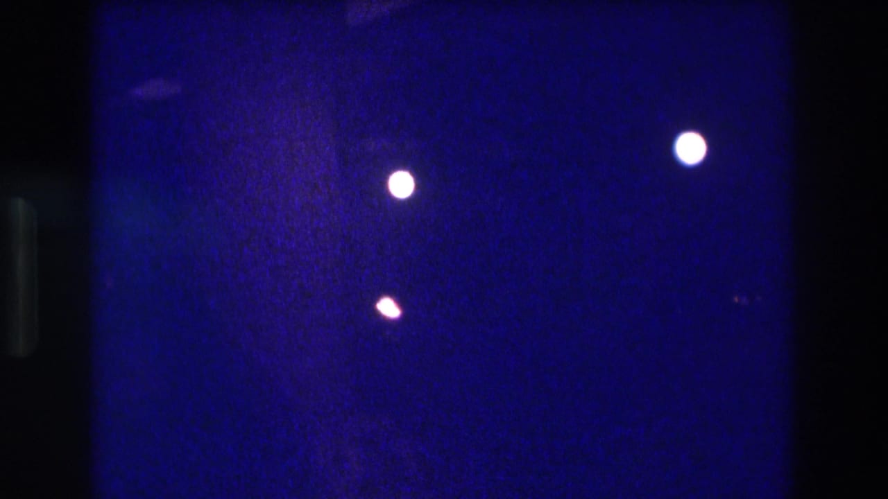 8mm frame from a circa-1982 Acapulco nighttime show, showing two white spotlight orbs and a smaller amber orb floating against a deep indigo field with visible film grain.