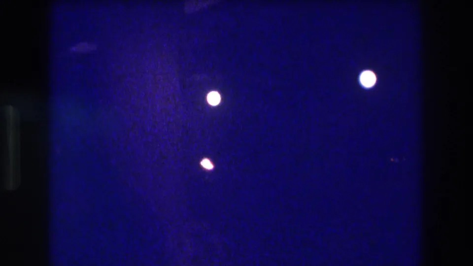 8mm frame from a circa-1982 Acapulco nighttime show, showing two white spotlight orbs and a smaller amber orb floating against a deep indigo field with visible film grain.