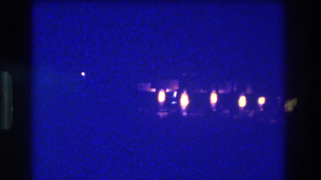 8mm home-movie frame circa 1982, Acapulco, Mexico — a row of warm amber-orange lights emerges along a distant waterfront against heavy blue-indigo grain.
