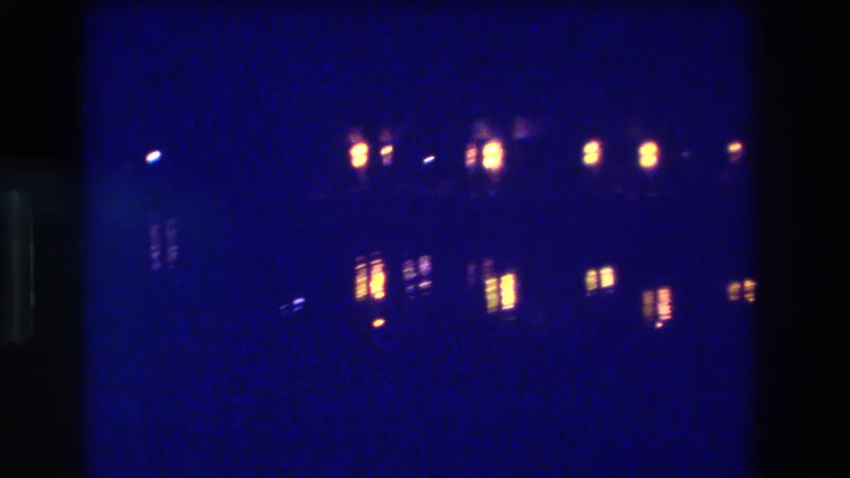 8mm home-movie frame circa 1982, Acapulco, Mexico — amber waterfront lights and their distinct shimmering reflections on dark water, rendered in deep blue grain.