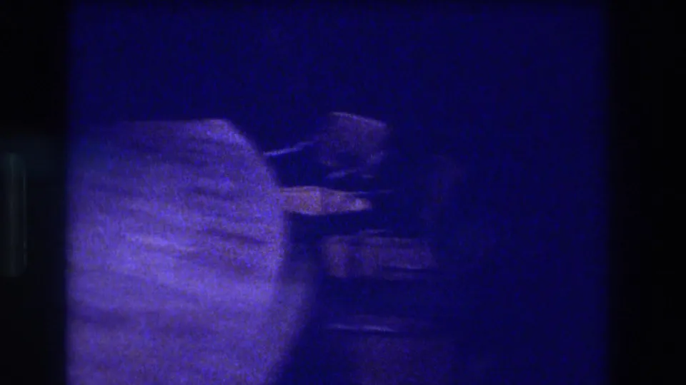 Extreme low-light 8mm interior shot, ca. 1982 Acapulco, deep blue cast and heavy grain obscuring a blurry foreground shape at left edge.