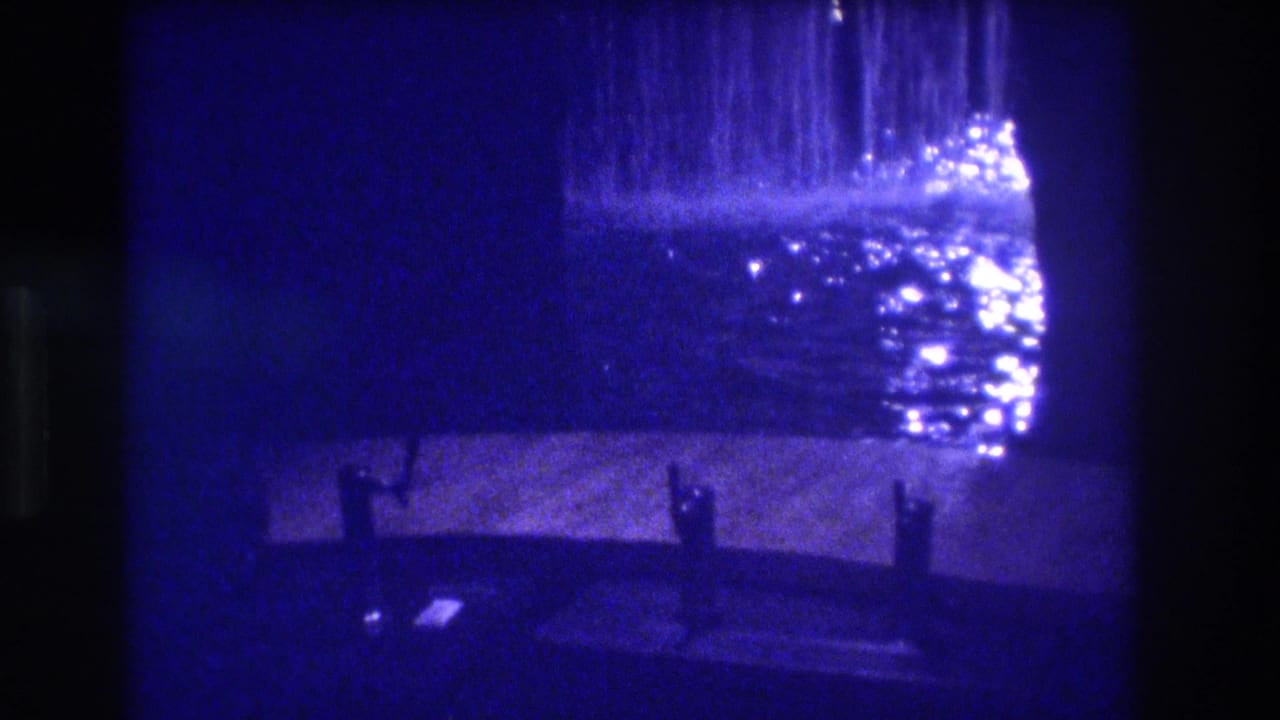 8mm home movie frame from Acapulco, 1982 — a cascading water feature lit from above, with dark silhouetted bollards or benches on wet pavement, deep blue film cast.