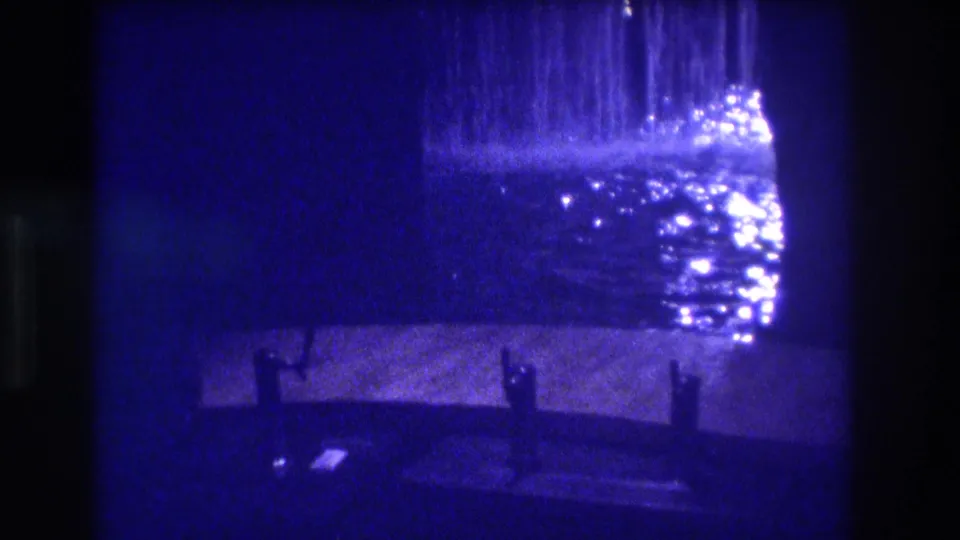 8mm home movie frame from Acapulco, 1982 — a cascading water feature lit from above, with dark silhouetted bollards or benches on wet pavement, deep blue film cast.