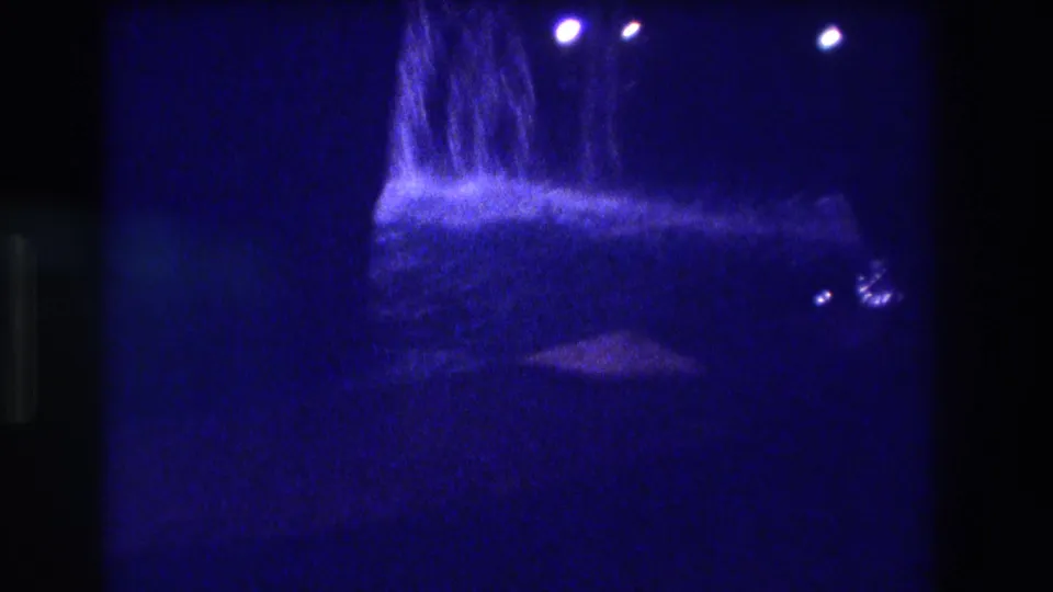 8mm home movie frame from Acapulco, 1982 — a broad waterfall or resort water feature with three lamp-style lights above it, dark canopy or tent shape at lower frame, heavy blue cast and grain.