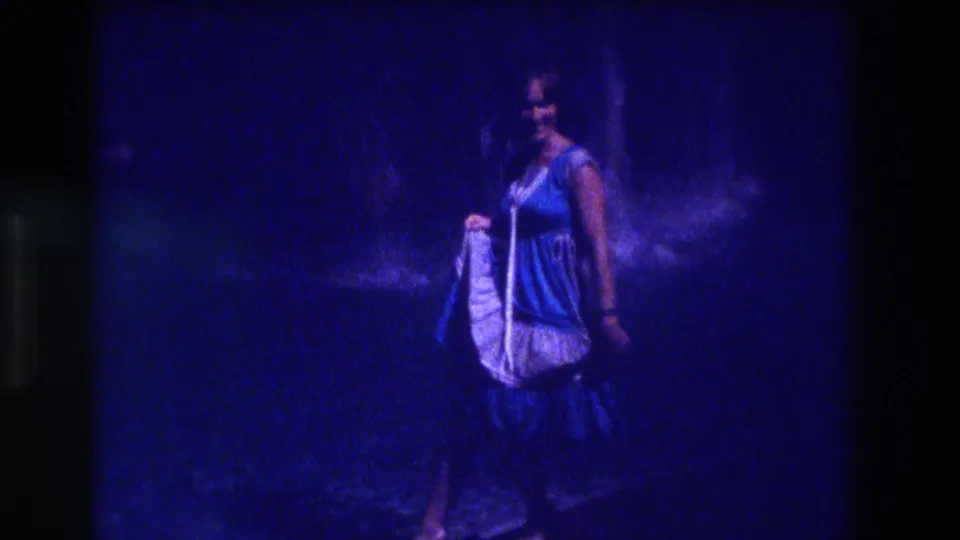8mm home movie frame from Acapulco, 1982 — a smiling adult woman in a light-colored sleeveless dress walks toward the camera at night, water cascade visible in background, strong blue film cast.