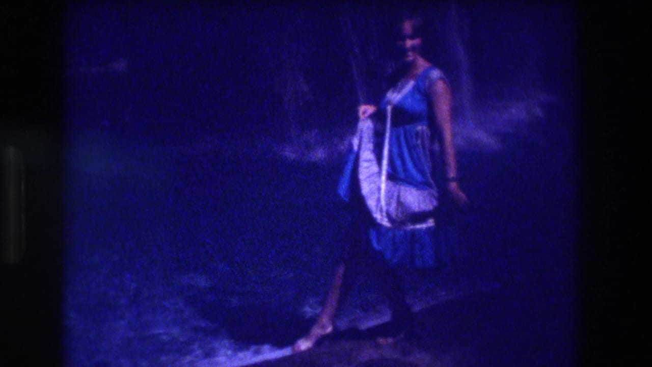 Woman in blue and white dress seen from the side, holding a white garment, against a dark nighttime background.