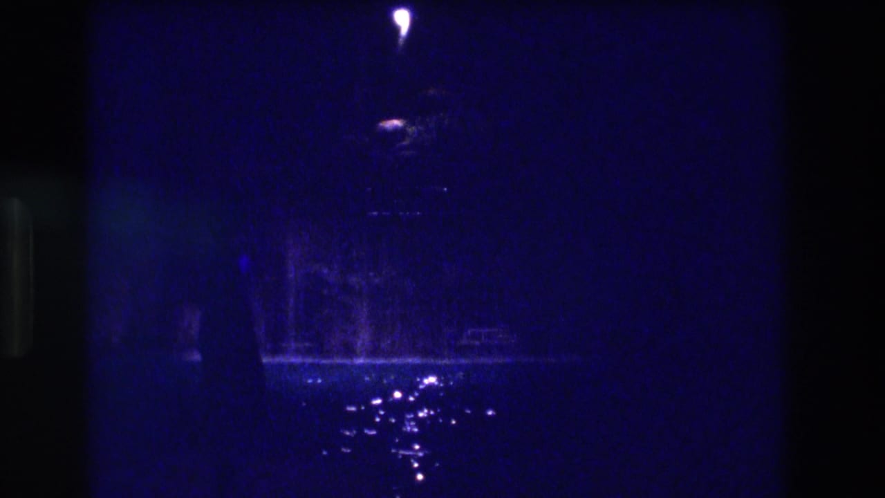 Nearly black 8mm frame: single bright point of light at top centre, circular lamp element below it, scattered reflections on wet ground