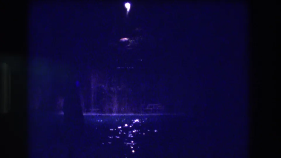 1982 Acapulco 8mm night frame showing a solitary point-light and scattered reflections on rain-wet pavement against a deep blue field