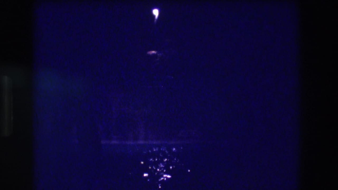 Second frame of apparent 1982 Acapulco night sequence — same solitary lamp point-light and rain-wet pavement reflections persist in deep blue