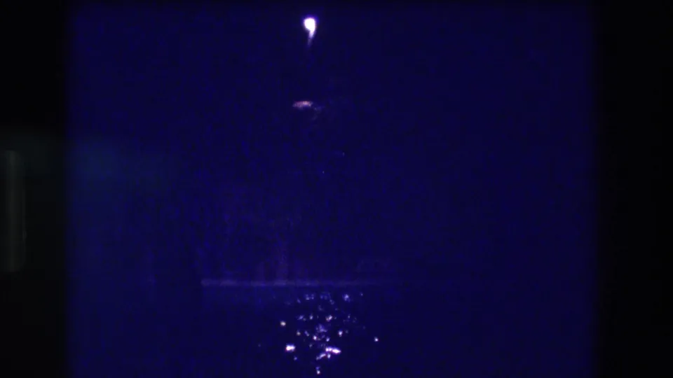 Second frame of apparent 1982 Acapulco night sequence — same solitary lamp point-light and rain-wet pavement reflections persist in deep blue