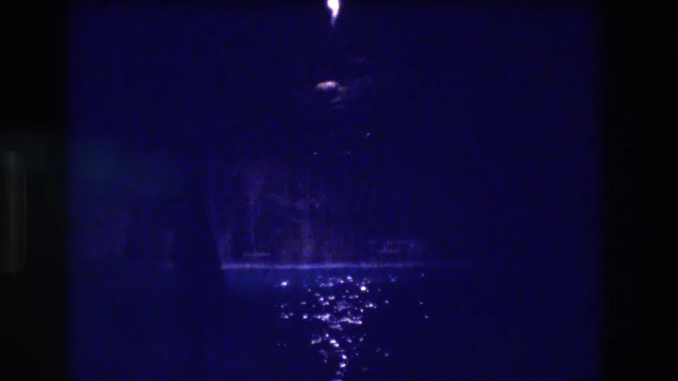 Mid-clip 8mm frame from apparent 1982 Acapulco night shoot — same lamp and wet-ground reflection composition with deep blue exposure and heavy grain