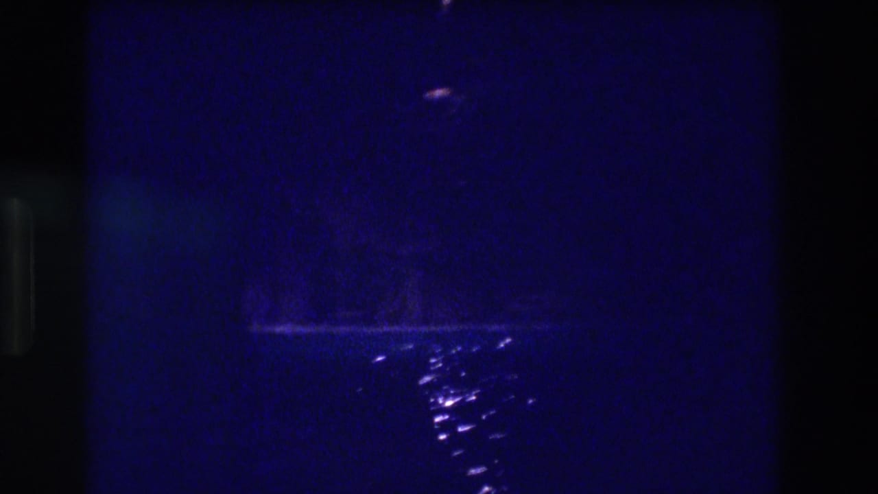 Deeply underexposed 8mm frame: blue darkness with a distant pinpoint light near the top and a diagonal streak of bright reflections on water below.