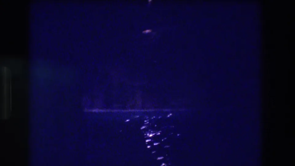 8mm home movie frame from 1982 Acapulco showing extreme blue-tinted darkness, a distant light near the top, and scattered water reflections on what may be a bay or ocean surface at night.