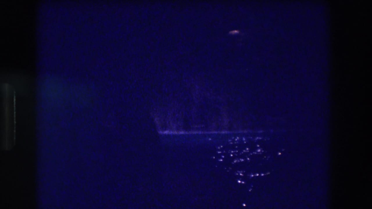8mm home movie frame from 1982 Acapulco at night: deep blue darkness, a distant reddish light near the top right, and a cluster of bright reflections on water below.
