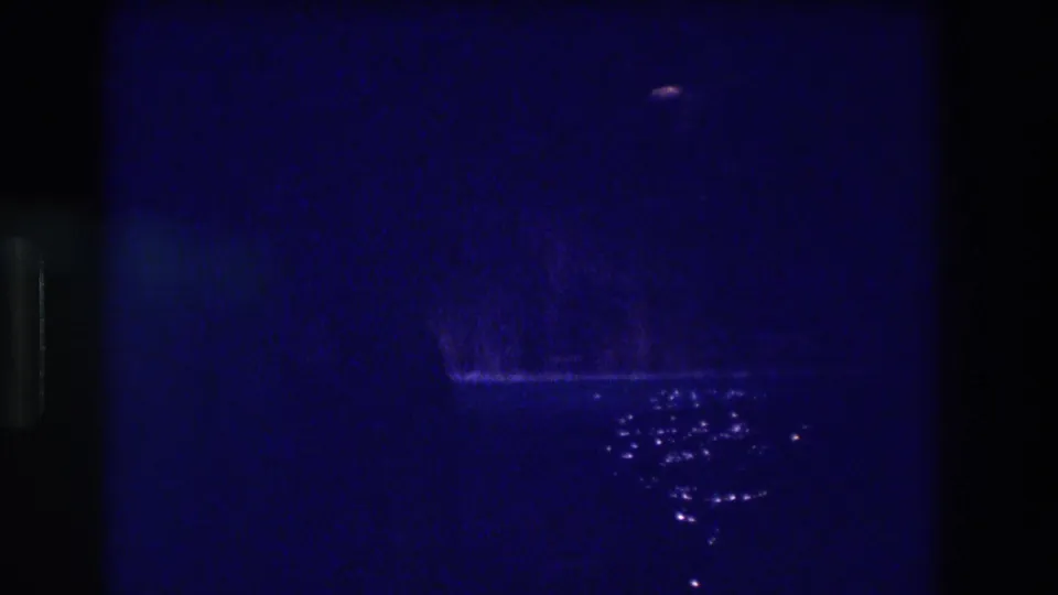 8mm home movie frame from 1982 Acapulco at night: deep blue darkness, a distant reddish light near the top right, and a cluster of bright reflections on water below.