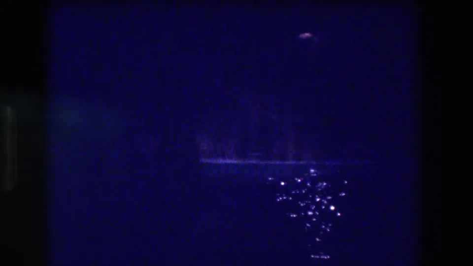 8mm home movie frame from 1982 Acapulco: deep blue darkness with a faint vertical lighter area, scattered water reflections, and film sprocket edge visible at left.