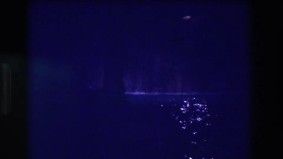 8mm home movie frame from 1982 Acapulco at night: blue-dominant darkness with a faint lit form or surface mid-left and scattered bright reflections on water.