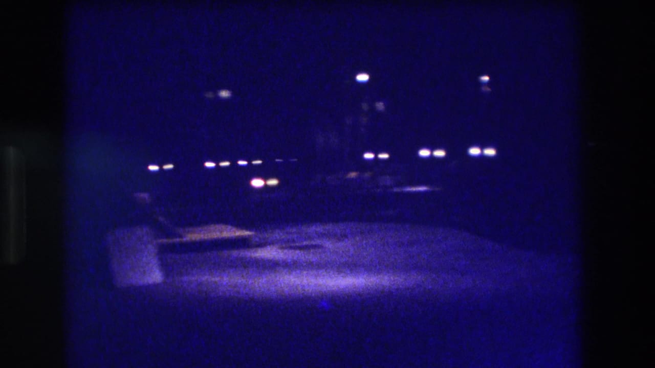 Deep blue night scene showing an open paved area with scattered distant light points and a low structure at left.