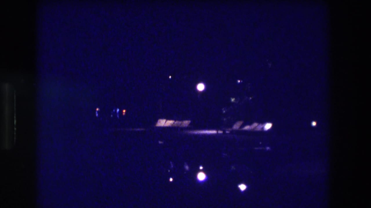 8mm home movie frame, Acapulco Mexico 1982 — dark coastal night with scattered shoreline lights and a bright horizontal band from what may be an elevated roadway or structure.